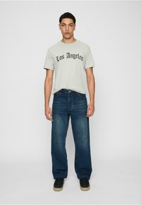Lys grå t-skjorte med "Los Angeles" i fet svart skrift, kombinert med løstsittende mørkeblå denimjeans og svarte sko med tan såle.