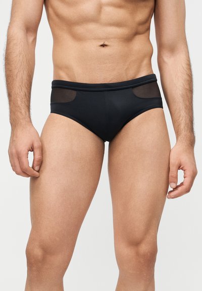 Ανδρικός κορμός και άνω μέρος των ποδιών που φορούν μαύρα εσώρουχα με διάφανα δίχτυα στα ισχία, σε απλό φόντο.