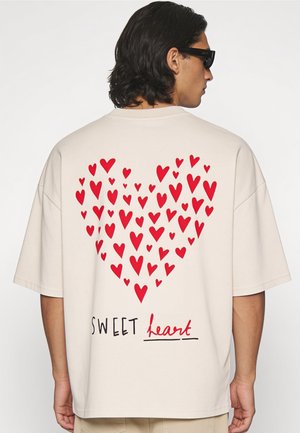 Homme aux cheveux noirs et lunettes de soleil, portant un t-shirt beige clair avec un motif de cœur rouge et le texte "SWEET heart" au dos.