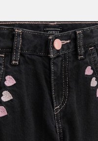 Schwarze Denim-Jeans mit bestickten rosa und hellrosa Herzen auf der Vorderseite. Kontrastnähte und ein gebrandeter Knopf an der Taille.