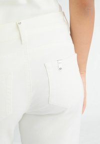 Weiße Denim-Jeans mit einer Nahaufnahme der hinteren Tasche, die ein silbernes Logoschild und eine subtile Textur zeigt. Einfacher Knopfverschluss an der Taille.