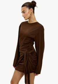 Robe marron à manches longues avec une silhouette ajustée, fronces sur le côté et un détail noué à la taille. Matière lisse et extensible avec une finition élégante.