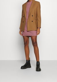Hnědý dvouřadový blazer přes růžové pletené šaty. Černé kotníkové boty se žlutým podpatek. Hladká textura a upravený střih.