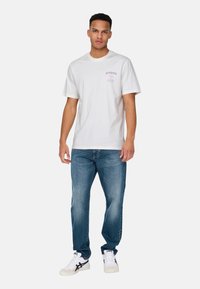 White cotton t-shirt with round neckline and short sleeves, featuring pink graphic text on the front, paired with blue denim jeans and white sneakers.