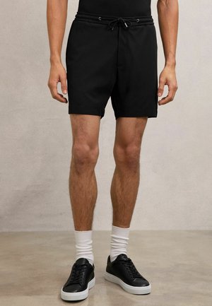 Moški, oblečen v črne kratke hlače z vrvico, bele nogavice in črne superge, stoji na betonski tlakovci ob svetlo sivi steni brez okraskov.