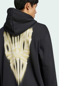 Juodas džemperis su dideliu geltonu grafiniu dizainu ir "Adidas" tekstu bei emblema ant nugaros. Pagamintas iš minkštos medžiagos su kapulėte.