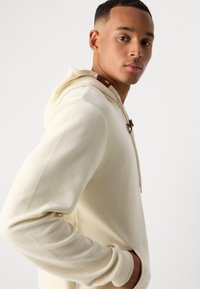 Junger Mann in einem cremefarbenen, gerippten Hoodie mit den Händen in den Taschen, der über die Schulter schaut, vor einem schlichten weißen Hintergrund.