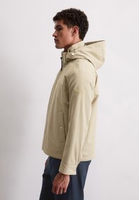 Beżowa kurtka z kapturem, zapinana na zamek błyskawiczny z kieszeniami na zatrzaski; charakteryzuje się gładką teksturą i regulowanym kapturem dla idealnego dopasowania.