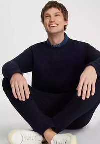 Ciemno granatowy sweatshirt i pasujące spodnie, wykonane z miękkiego materiału, nosi siedzący mężczyzna. Białe sneakersy z teksturowanymi podeszwami dopełniają strój.