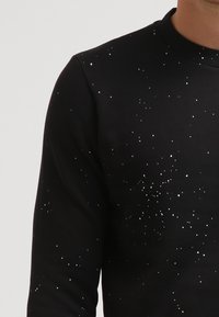 Maglietta nera a maniche lunghe con un piccolo motivo di macchie bianche che somigliano a stelle sulla parte superiore del corpo e sul braccio sinistro.