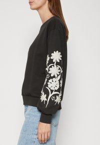 Femme portant un sweat-shirt noir à manches longues avec broderies florales blanches sur les manches et un jean bleu clair, vue de profil.