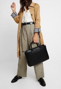 Kvinde iført en tan trench coat med ternede manchetter, hvid skjorte, beige vide bukser, sort bælte, sorte loafers og bærende en sort håndtaske.