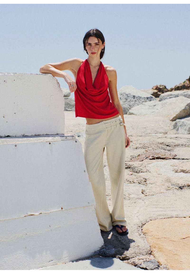Femme en haut dos nu rouge et pantalon beige appuyée sur un bloc de béton blanc en extérieur sur un sol rocheux sous un ciel dégagé.