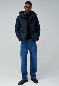 Veste à capuche bleu marine avec poches latérales, portée par-dessus un pull noir. Jean bleu clair avec une coupe droite, associé à des bottes noires.