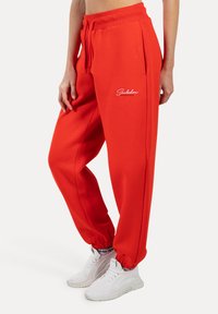Rode sweatpants met een koord in de taille, zijzakken en elastische manchetten. Heeft een wit logo op de linkerdij. Draagt het met witte sneakers.