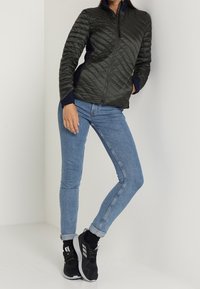 Mörkgrön vadderad jacka med en figurnära design, kombinerad med ljusblå skinny jeans och svarta sportskor med vita detaljer.