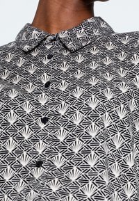 Czarna i biała koszula w wzory, z geometrycznym wzornictwem, kołnierzykiem oraz sześcioma czarnymi guzikami z przodu, o gładkiej fakturze materiału.