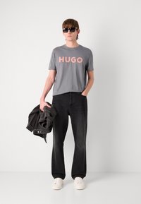 Pilkas marškinėliai su rožiniu "HUGO" logotipu, juodos džinsinės kelnės, laikantis juodą džinsinį švarką ir dėvint juodus akinius nuo saulės. Balti sportbačiai užbaigia įvaizdį.