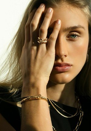 Guld ringe og et struktureret guldarmbånd pryder en hånd, der dækker en del af ansigtet, med en neutral baggrund, der fremhæver smykkernes design.