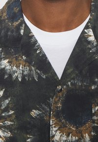 Camicia a maniche corte con un motivo floreale in verde scuro, oro e bianco. Chiusura con bottoni, dettaglio del colletto e indossata sopra una maglietta bianca.