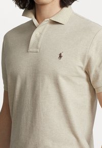 Beżowa koszulka polo z teksturowanej tkaniny, z trzema guzikami i małym brązowym haftowanym logo na lewej piersi. Krótkie rękawy.