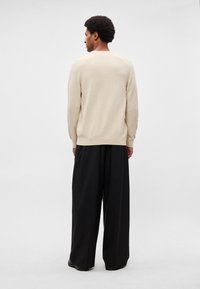 Suéter beige de punto con puños y dobladillo acanalados, combinado con pantalones negros de corte ancho, visto desde atrás sobre un fondo claro.