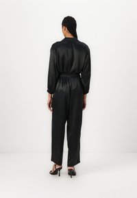 Mustast satiinist jumpsuit, millel on pikad varrukad, painduva vöökoha ja laiad sääred. Kantud mustade rihmadega kontsakingadega.