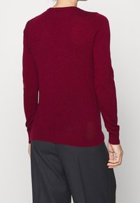 Burgunderfarbenes Langarmshirt mit Rundhalsausschnitt, weichem Material, taillierter Form und gerippten Abschlüssen, getragen mit schwarzen weiten Hosen.