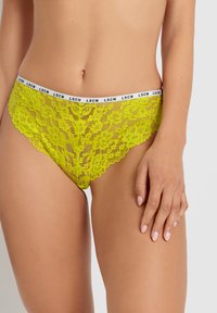 Koronkowe majtki bikini w fluorescencyjnym żółtym kolorze z kwiatowym wzorem, wyposażone w czarno-białą elastyczną talię z napisem "LSCN."