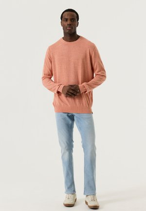 Homme debout portant un pull corail à manches longues, un jean bleu clair et des baskets blanches, sur un fond clair uni.