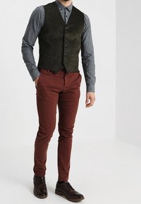 Gilet in corda verde scuro sopra una camicia grigia con bottoni, abbinato a pantaloni slim-fit color ruggine e scarpe stringate in pelle marrone.