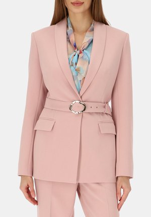 Kobieta w pastelowo różowym blezerze z paskiem i klapowymi kieszeniami, nosząca pod spodem niebieską i różową bluzkę we florystyczny wzór z wiązaniem przy dekolcie.