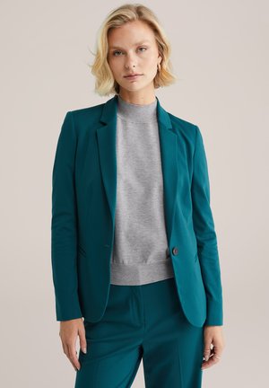 Blazer teal con un diseño entallado, cierre de un botón y solapas medianas, llevado sobre un suéter de cuello alto gris claro, combinado con pantalones a juego.