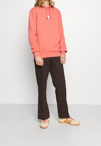 Korálová mikina s kapucí a přední kapsou, s kapucí na stažení, kombinovaná s tmavě hnědými kalhotami a béžovými botami s oranžovými pruhy.