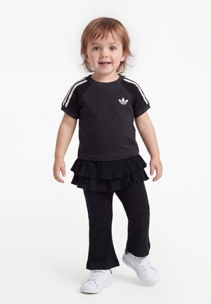 Νήπιο που φοράει μαύρο μπλουζάκι Adidas με λευκές ρίγες, μαύρη πολυεπίπεδη φούστα πάνω από παντελόνια και λευκά αθλητικά παπούτσια, στέκεται σε λευκό φόντο.