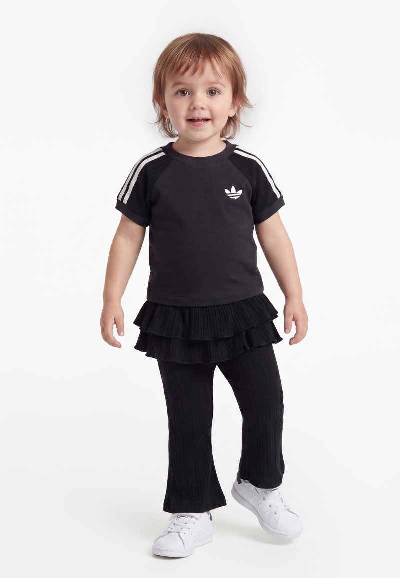 Νήπιο που φοράει μαύρο μπλουζάκι Adidas με λευκές ρίγες, μαύρη πολυεπίπεδη φούστα πάνω από παντελόνια και λευκά αθλητικά παπούτσια, στέκεται σε λευκό φόντο.