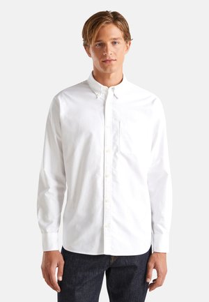 Camisa blanca de manga larga con botones que presenta un cuello, un bolsillo en el pecho y una textura de algodón suave. Combinada con jeans oscuros de mezclilla.