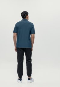 Polo shirt corto in verde scuro con colletto, texture liscia e dettagli sottili sulla schiena, abbinato a pantaloni neri tapered e sneakers bianche.