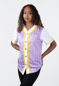 Maglia a righe viola con accenti gialli, maniche bianche e chiusura a bottoni. Presenta la scritta "KANI" sul davanti. Tessuto liscio.