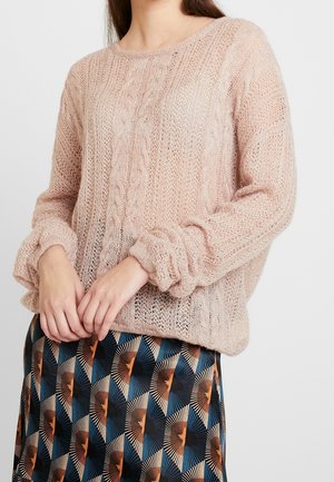 Femme portant un pull tricoté ample rose pâle associé à une jupe colorée à motifs géométriques avec des formes bleues, noires et orange.