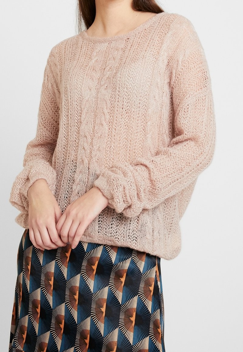 Femme portant un pull tricoté ample rose pâle associé à une jupe colorée à motifs géométriques avec des formes bleues, noires et orange.