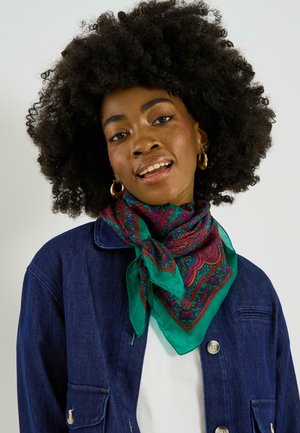 Femme aux grands cheveux bouclés, portant des boucles d'oreilles anneaux dorés, une écharpe colorée à motifs, une chemise blanche et une veste bleu foncé.