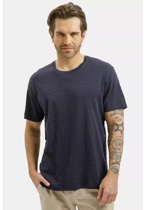 Mężczyzna z zaczesanymi do tyłu włosami, ubrany w granatowo paskowaną koszulkę i beżowe spodnie, pokazujący tatuaże na obu ramionach, stoi z jedną ręką w kieszeni.