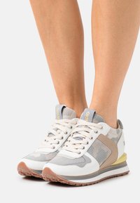 Baskets blanches et grises avec des accents en maille et en daim, bout rond, accents beiges, semelle en caoutchouc, et un détail jaune sur la languette du talon.