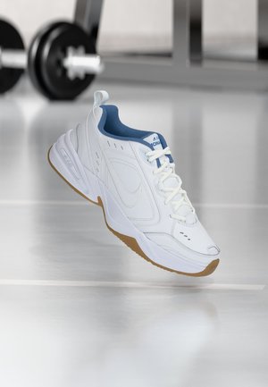 Fehér atlétikai cipő kék belsővel és gumitalppal, lebeg egy tükröződő terem padló felett, elmosódott súlyzórúddal a háttérben.