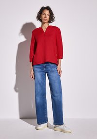 Blusa rossa di lino con maniche a tre quarti e scollatura a V, abbinata a jeans in denim blu larghi e sneakers bianche, su uno sfondo neutro.
