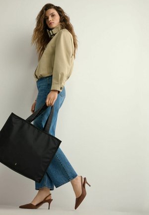 Kobieta ubrana w beżową kurtkę i niebieskie dżinsy, trzymająca dużą czarną torbę typu tote i mająca na stopach brązowe sandały na obcasie, pozująca na tle gładkiej ściany.