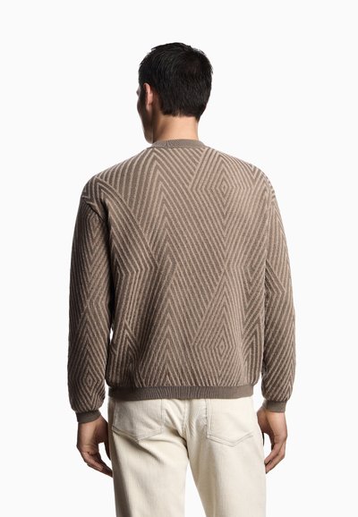 Maglione marrone con motivo geometrico, realizzato in tessuto testurizzato, con polsini e orlo a coste. Indossato sopra pantaloni di velluto a coste color avorio.