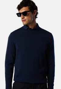 Maglione lavorato a maglia blu navy con scollatura rotonda e maniche lunghe, realizzato in un materiale liscio e testurizzato. Indossato sopra una camicia con colletto.
