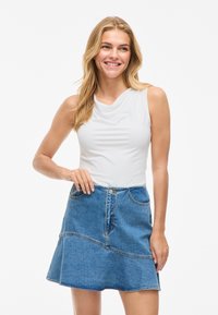 Smiling woman with long blonde hair wearing sleeveless white top and blue denim skirt, standing against white background.